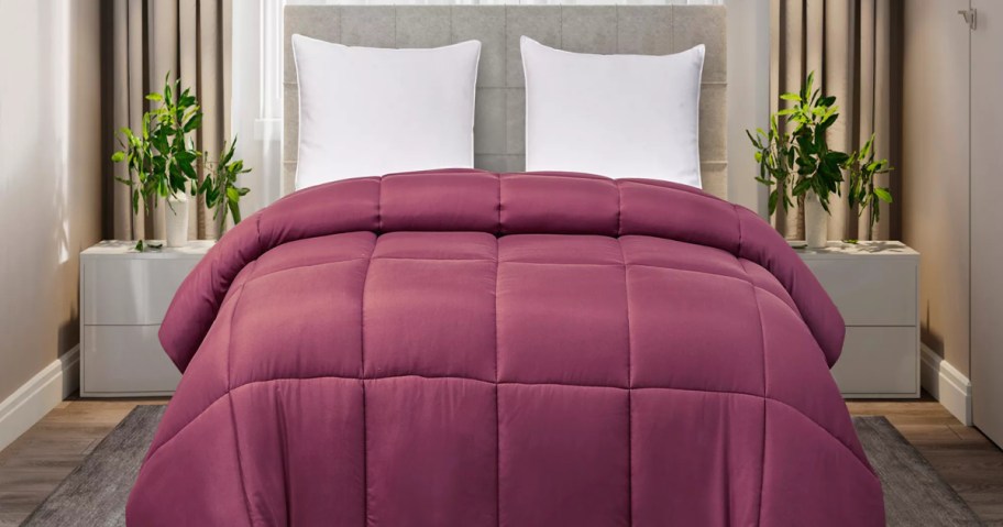 A bed made with a dark mauve or purple quilted down alternative comforter and two white pillows against a neutral headboard in a bedroom.
