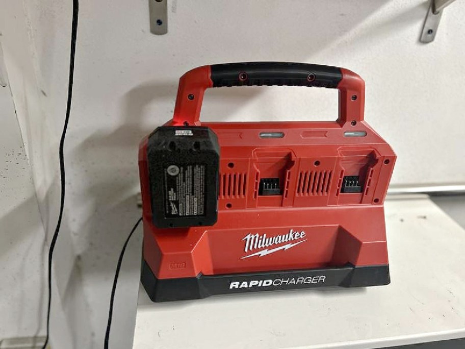 A red Milwaukee PACKOUT 6-port rapid charger with an attached battery resting on a white shelf.