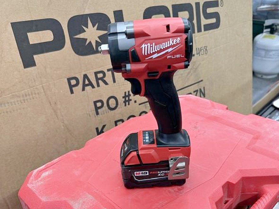 A Milwaukee M18 FUEL cordless impact wrench with an attached battery standing upright on a red surface in front of a cardboard box.