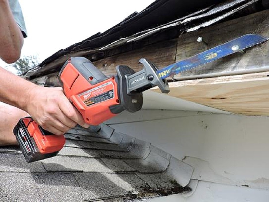 A person using a Milwaukee cordless reciprocating saw to cut into wooden roof eaves near shingles.
