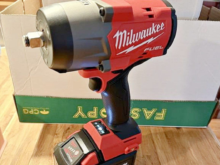 A Milwaukee M18 FUEL mid-torque impact wrench with an attached battery resting on a wooden surface in front of a box.