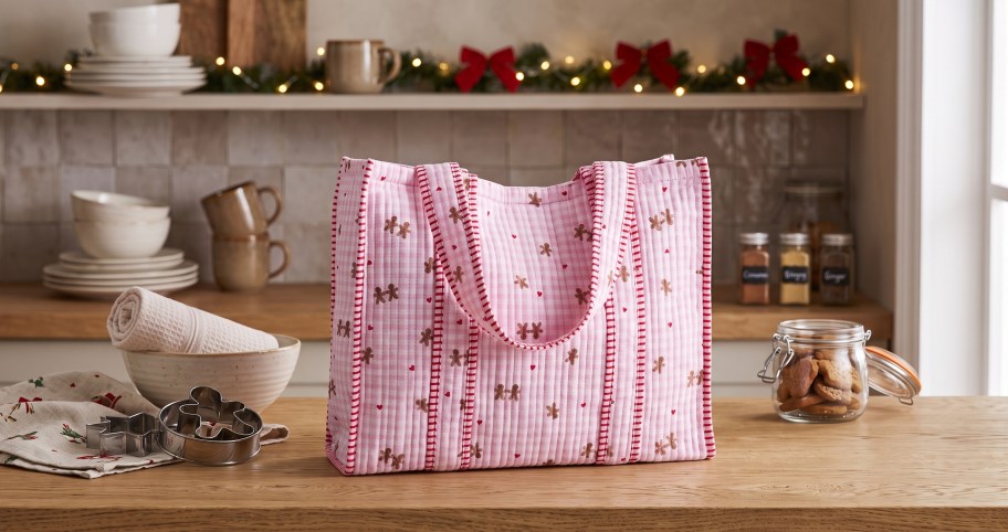 Pink and red patterned quilted tote bag lying flat on a wooden surface next to metal cookie cutters and a glass jar of cookies.