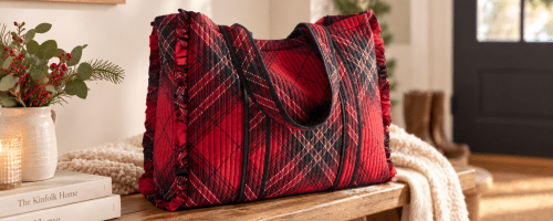 Red and black plaid quilted tote bag with ruffled sides resting on a wooden bench in a cozy entryway with books, greenery, and a cream throw blanket nearby.