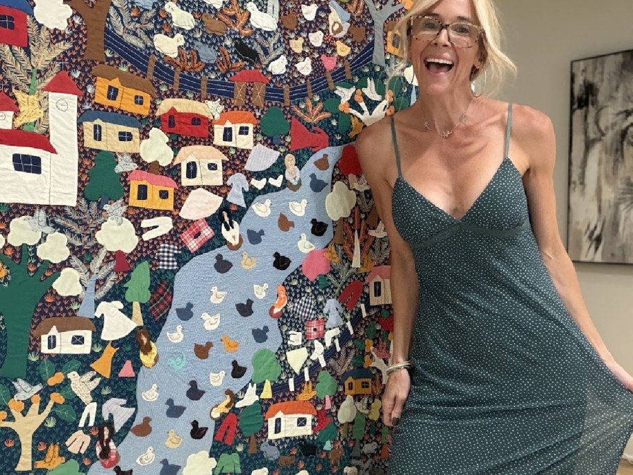 A person wearing a dark green polka-dot maxi dress standing in front of a colorful patterned wall hanging.