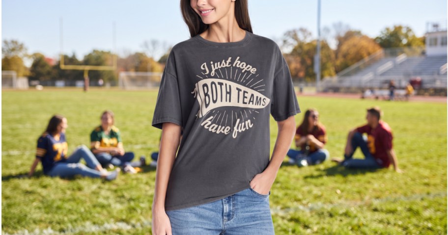A woman wearing a charcoal grey graphic t-shirt that says "I just hope both teams have fun" with jeans while standing outdoors on a sports field.