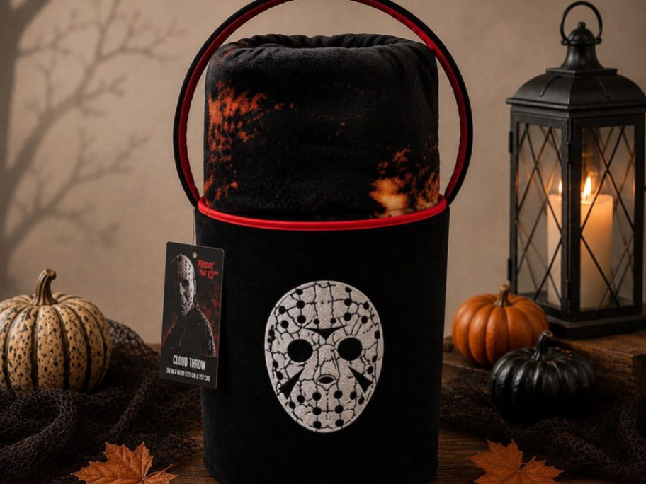 A Friday the 13th Halloween throw blanket and storage bucket set featuring Jason's hockey mask on the front, flanked by small pumpkins and a lit lantern.