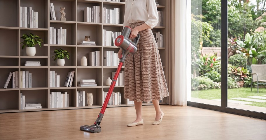 Woman vacuuming a light hardwood floor using a red and grey cordless stick vacuum in a modern living room with floor-to-ceiling bookshelves.