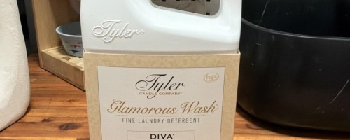 A large jug of Tyler Glamorous Wash fine laundry detergent in the Diva scent set on a wooden surface.