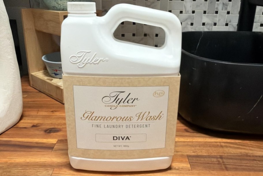 A large jug of Tyler Glamorous Wash fine laundry detergent in the Diva scent set on a wooden surface.