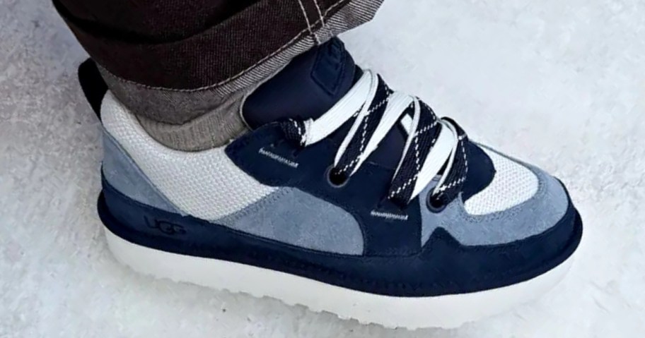 A close-up of a person wearing UGG Men's Lo Lowmel shoes with white and black patterned laces, dark blue and gray panels, and a thick white platform sole.