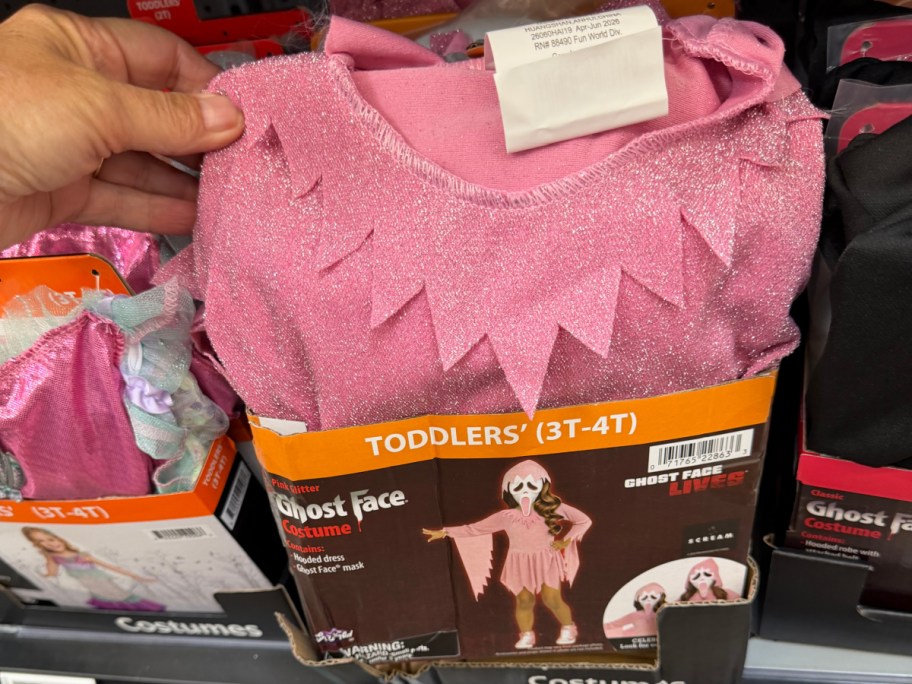 A hand holding a toddler-sized pink glitter Ghost Face costume box on a store shelf.