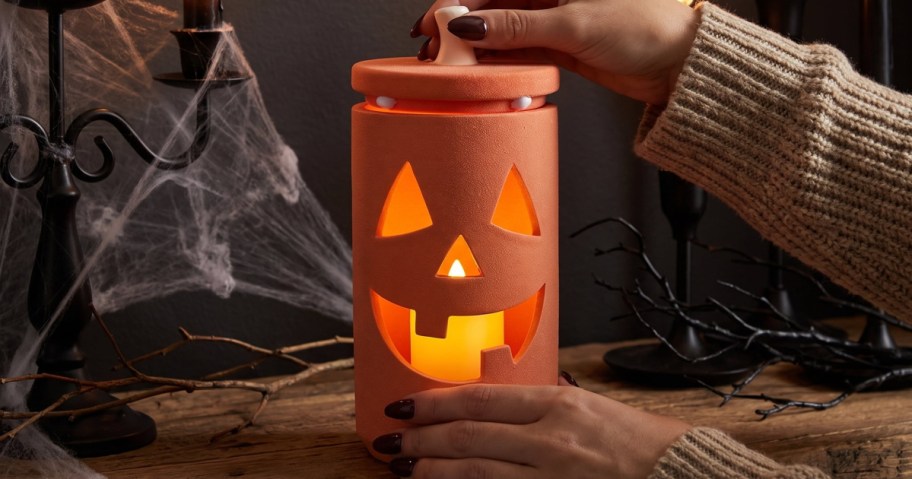 A pair of hands lifting the lid off a cylindrical orange ceramic jack-o'-lantern jar with a lit candle inside, set against a Halloween-themed background with cobwebs and candlesticks.