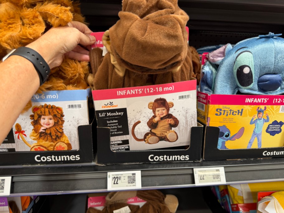A person's hand holding a Way to Celebrate infant Lil' Monkey Halloween costume box on a store shelf, flanked by a lion costume on the left and a Disney Stitch costume on the right.