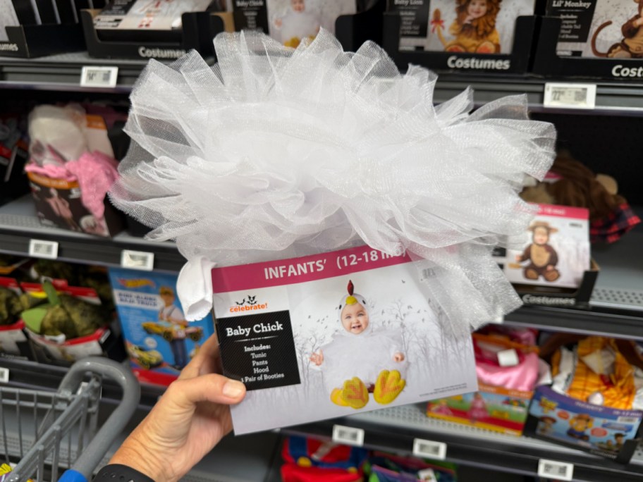 A hand holding a Way to Celebrate infant Baby Chick Halloween costume box topped with a large white fluffy pouf on a store shelf.