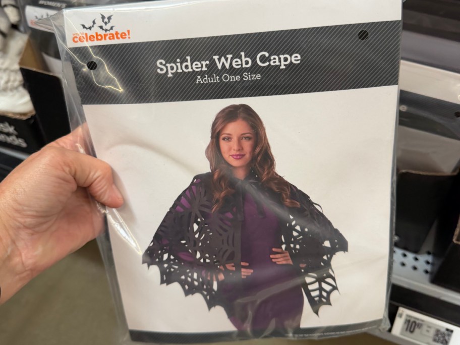 A hand holding a Way to Celebrate Spider Web Cape adult costume accessory package on a store shelf.