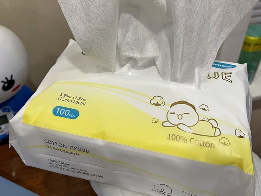 A pack of 100% cotton dry wipes with a yellow and white package featuring a cartoon baby graphic and tissue pulling out of the top.