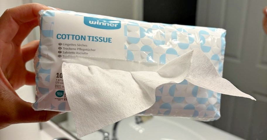 A hand holding a package of Winner cotton tissues with one tissue pulled out from the dispenser opening.