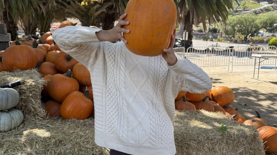 a woman in a white oversized cable knit sweater with a pumpkin in front of her face