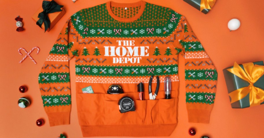 An orange and green Home Depot holiday sweater featuring festive tool patterns and pockets holding hardware tools, laid out against an orange background with wrapped gifts and ornaments.