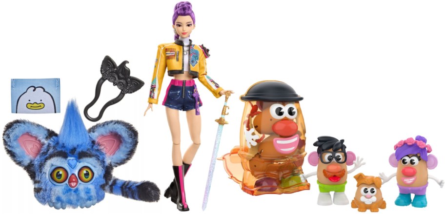 A collection of toys including a blue furry furby plush on the left, a kpop demon hunters fashion doll with purple hair holding a sword in the center, and several potato-head toy figures and accessories on the right.