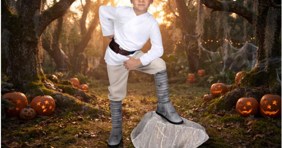 A child wearing a Luke Skywalker costume standing in a forest with carved pumpkins.