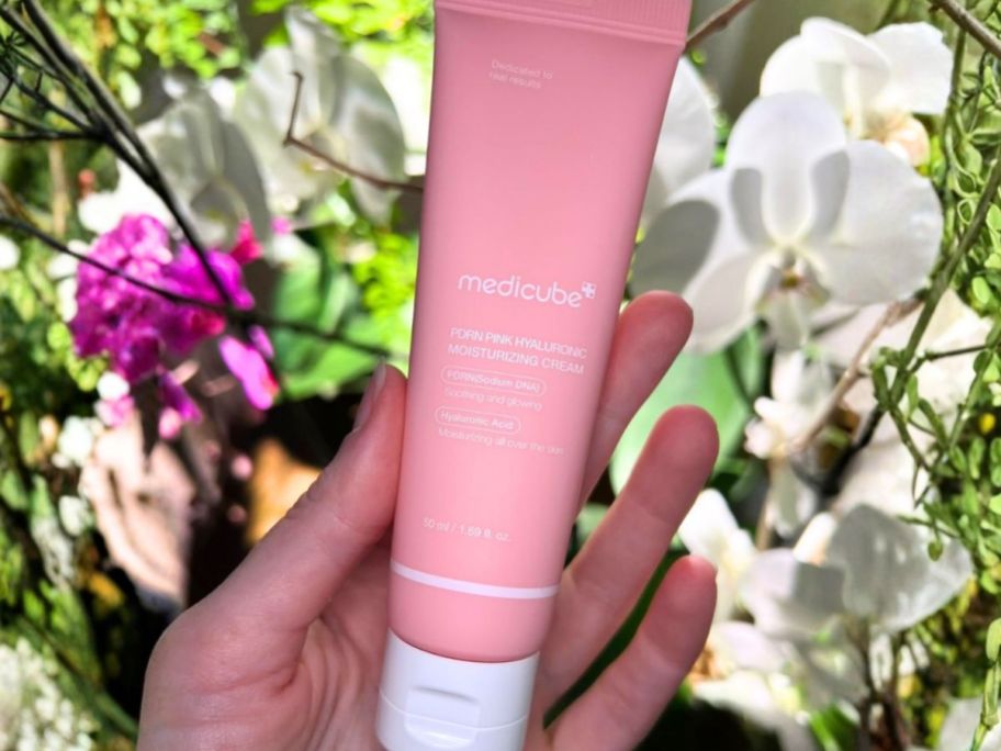 hand holding medicube Salmon DNA PDRN Pink Hyaluronic Moisturizing Cream tube in front of flowers