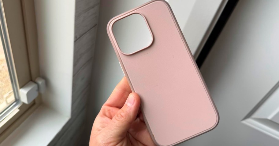 A hand holding a solid pink OtterBox phone case indoors near a window.