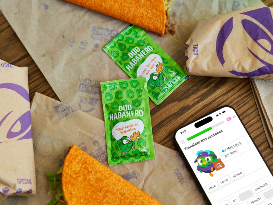 A spread of Taco Bell food packaging, Duo Habanero sauce packets, and a smartphone displaying a Duolingo translation lesson.