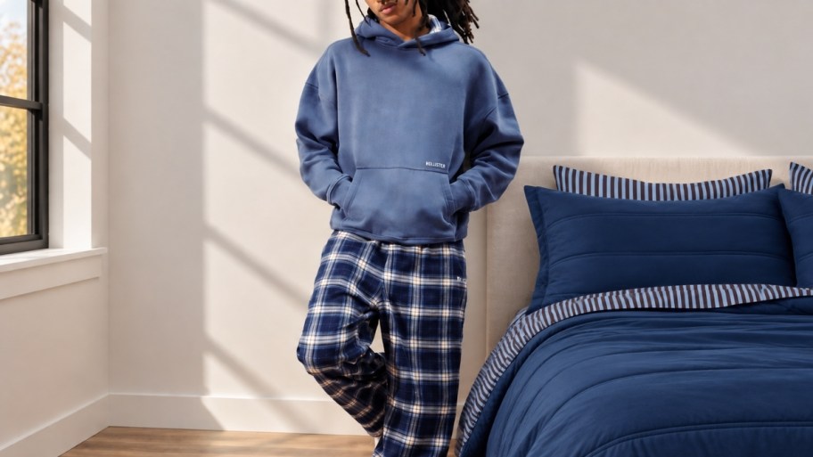 a man in a blue hoodie and plaid pj pants leaning against a wall