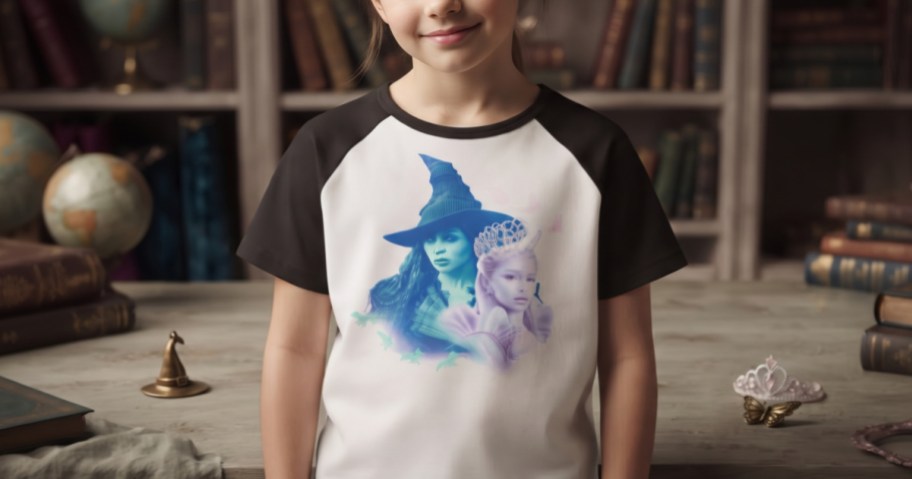 A young girl wearing a white and black raglan t-shirt with a Wicked movie graphic, standing in front of a bookshelf.