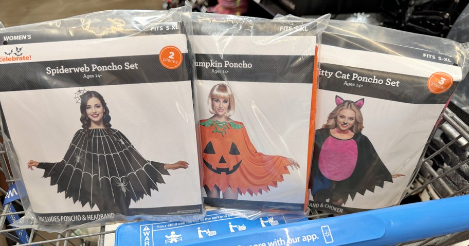 Three Way to Celebrate Halloween costume packages displayed side by side in a shopping cart, featuring a spiderweb poncho set, a pumpkin poncho, and a kitty cat poncho set.
