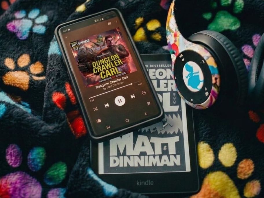 A phone and a kindle displaying Dungeon Crawler Carl on the screens and a pair of headphones next to them