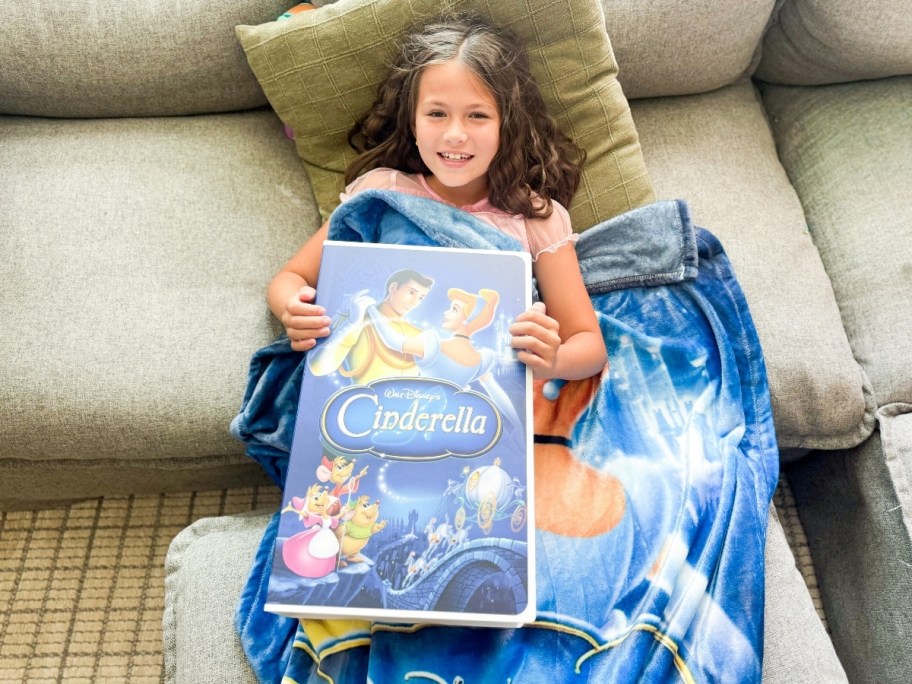 girl holding vhs style box with cinderella movie cover while wearing blanket on couch