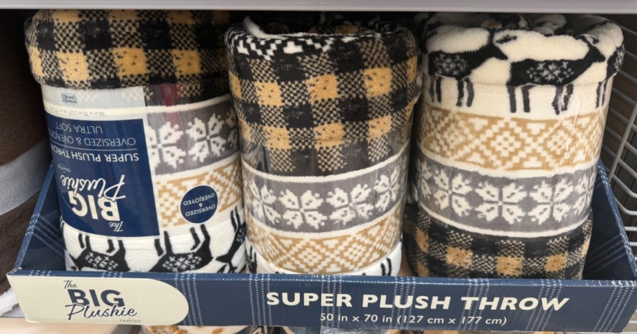 Three Crafted by Catherine The Big Plushie super plush throw blankets with holiday patterns, plaid, and reindeer designs displayed in a store box.