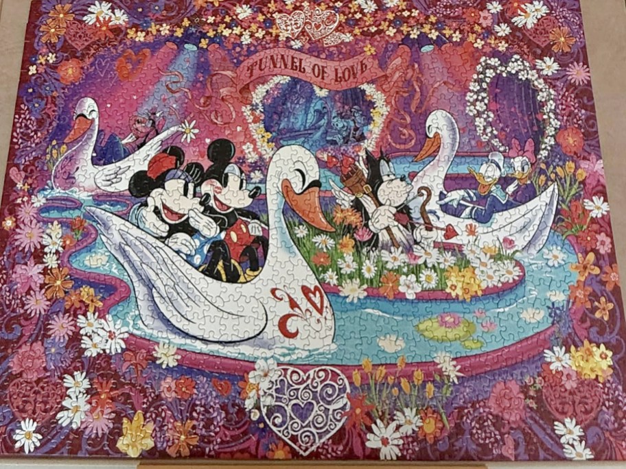 A completed 1000-piece Disney "Tunnel of Love" jigsaw puzzle featuring Mickey and Minnie Mouse riding in a swan boat surrounded by flowers and other Disney characters.