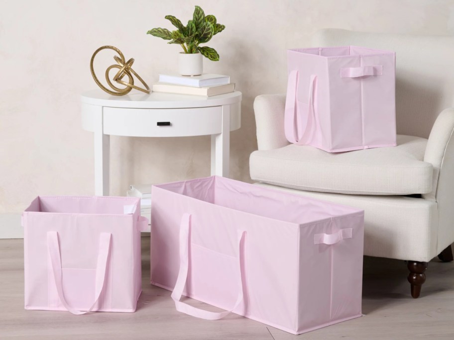 A set of three collapsible pink storage totes with carrying handles displayed in a room with a white armchair and side table.