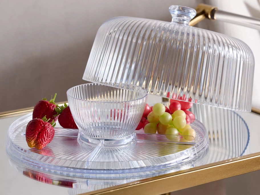 A clear, ribbed acrylic 3-in-1 serving set configured with a divided platter, a matching small bowl in the center, and a domed lid tilted open, filled with fresh strawberries and grapes.