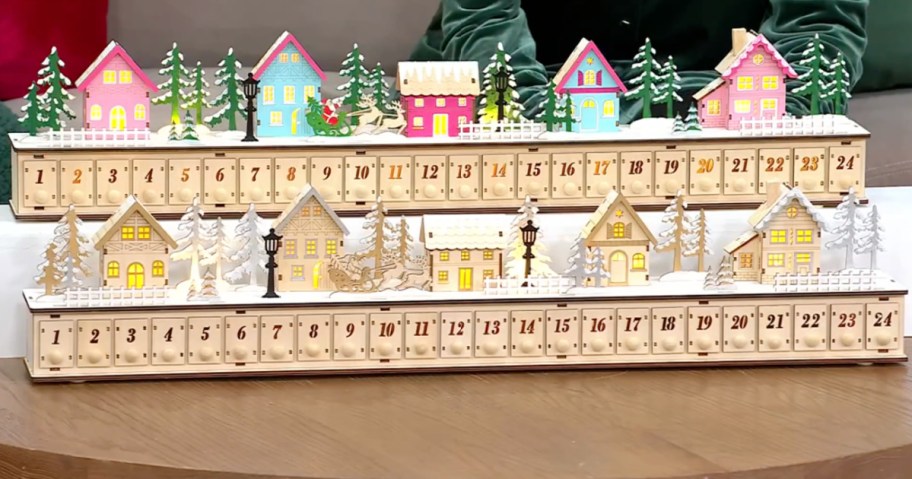 Two tiered wooden Home Reflections Christmas village advent calendars displayed on a table, featuring numbered countdown drawers numbered 1 through 24 beneath illuminated holiday village scenes with miniature houses and trees.