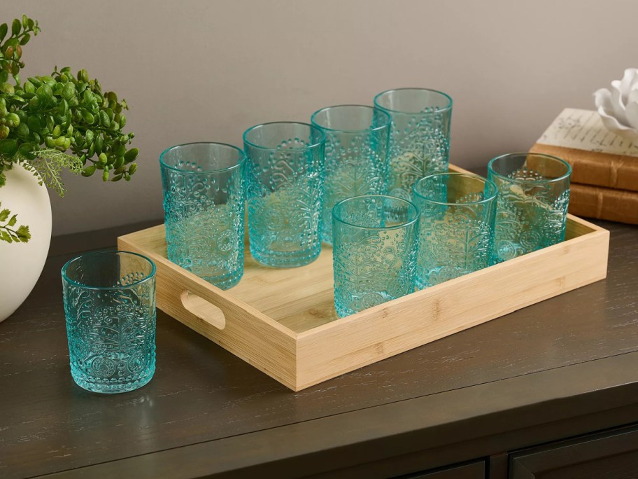 A set of eight embossed teal glassware tumblers displayed on a wooden serving tray on a tabletop.