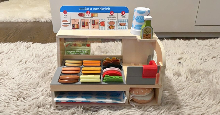 Melissa & Doug wooden sandwich play set featuring a counter with toy ingredients, a pretend cash register, a menu board, and accessories on a fluffy rug.