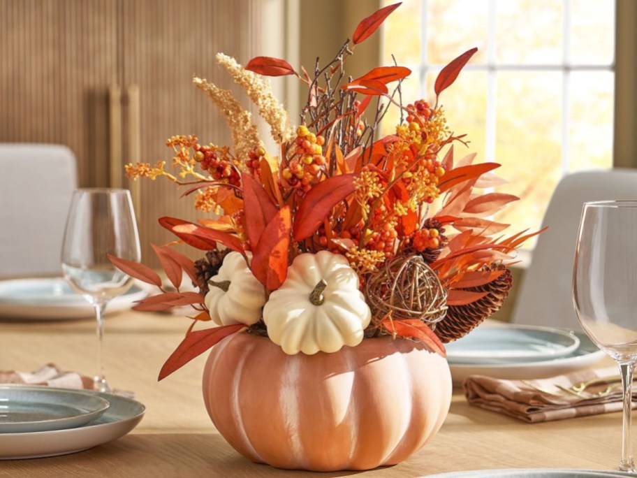 A Member's Mark 15-inch harvest terra cotta floral arrangement featuring white pumpkins, fall leaves, berries, and pinecones arranged inside a terra cotta pumpkin planter on a dining table.