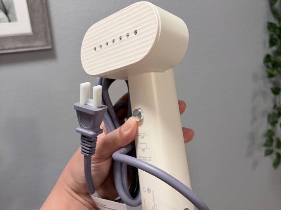 Nexdoo handheld travel clothes steamer in white with a gray power cord and plug being held up.