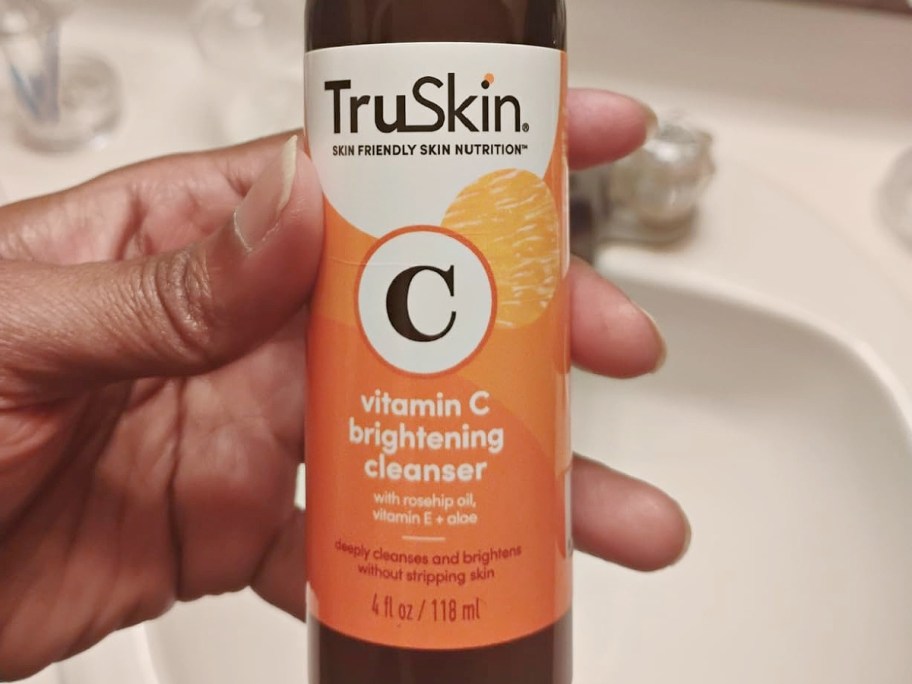 A bottle of TruSkin Vitamin C brightening cleanser held in a hand over a bathroom sink.