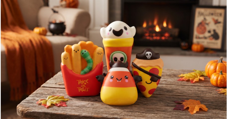 A collection of Way to Celebrate Spooky Snacks mystery Halloween squishy toys arranged on a wooden table with fall leaves, featuring french fries with a worm, a ghost drink cup, a bat-winged candy corn, and a pirate pizza slice.