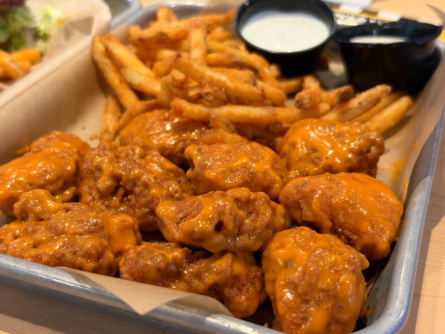A metal tray lined with paper holding buffalo wings coated in orange sauce, french fries, and cups of dipping sauce.