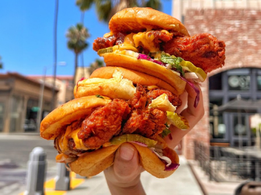 A hand holding a stack of three Dave's Hot Chicken sliders outdoors against an urban street background.
