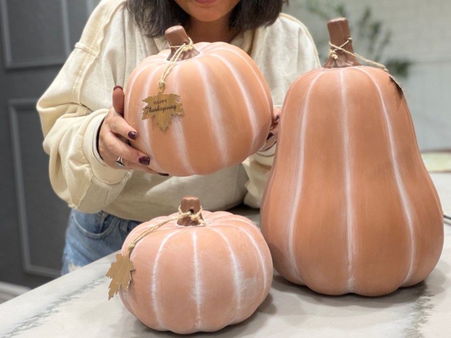 A person holding a light-colored terra cotta pumpkin with white accent lines and a Happy Thanksgiving tag, with two additional terra cotta pumpkins sitting on the counter.