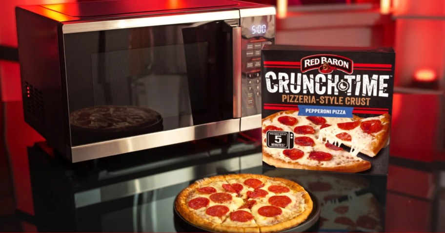 A cooked pepperoni pizza on a tray next to a Red Baron Crunch Time pizza box and a microwave on a dark surface.