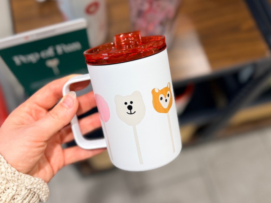A person holding a white Starbucks travel mug with animal cake pop illustrations and a red lid.