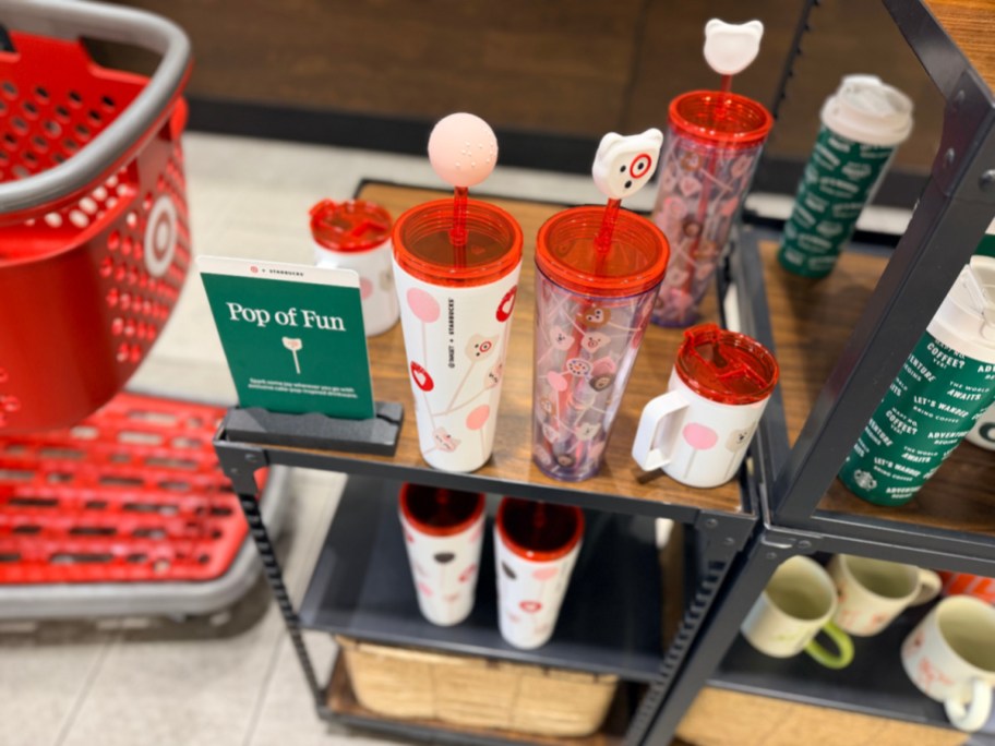 Starbucks "Pop of Fun" cake pop-inspired drinkware collection featuring tumblers, mugs, and a display sign on a store shelf next to a red shopping cart.